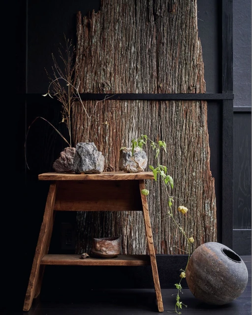 Still life composition featuring a rustic wooden stool, stone planters with greenery, and a textured wooden backdrop against a dark wall.