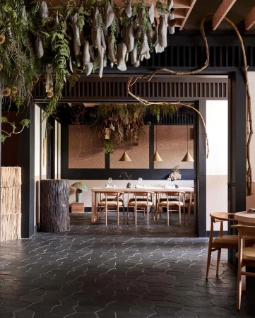 Dining room interior with a long wooden table, woven chairs, dark flooring, and hanging botanical installations above.