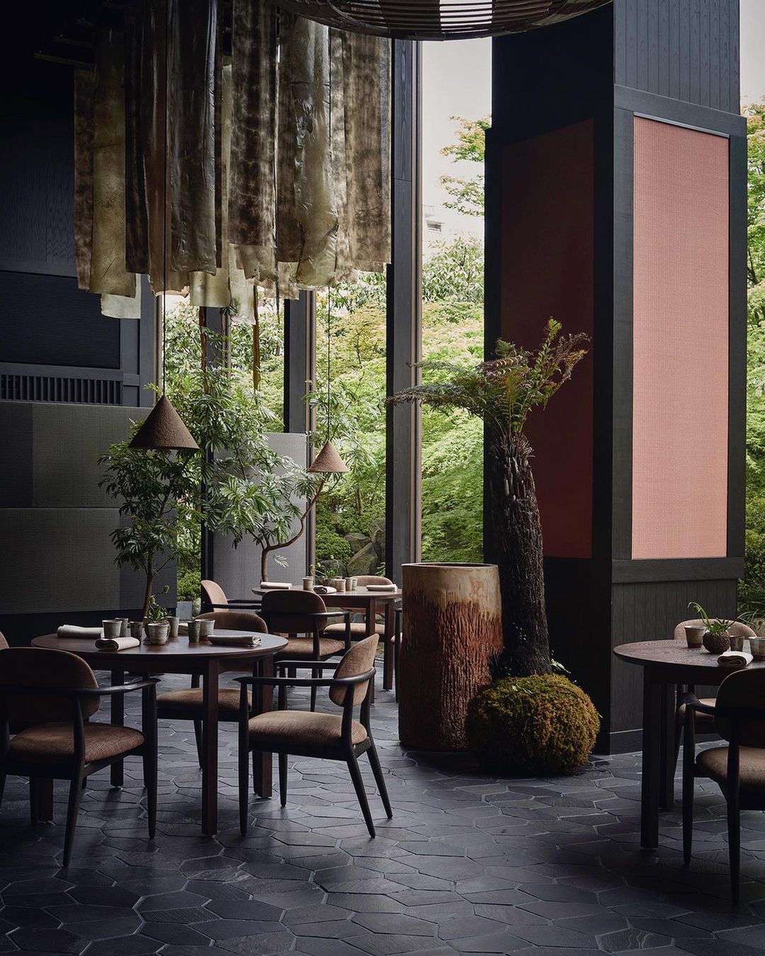 An elegant restaurant interior features tables set for dining, illuminated by pendant lights, with a backdrop of lush greenery seen through windows.
