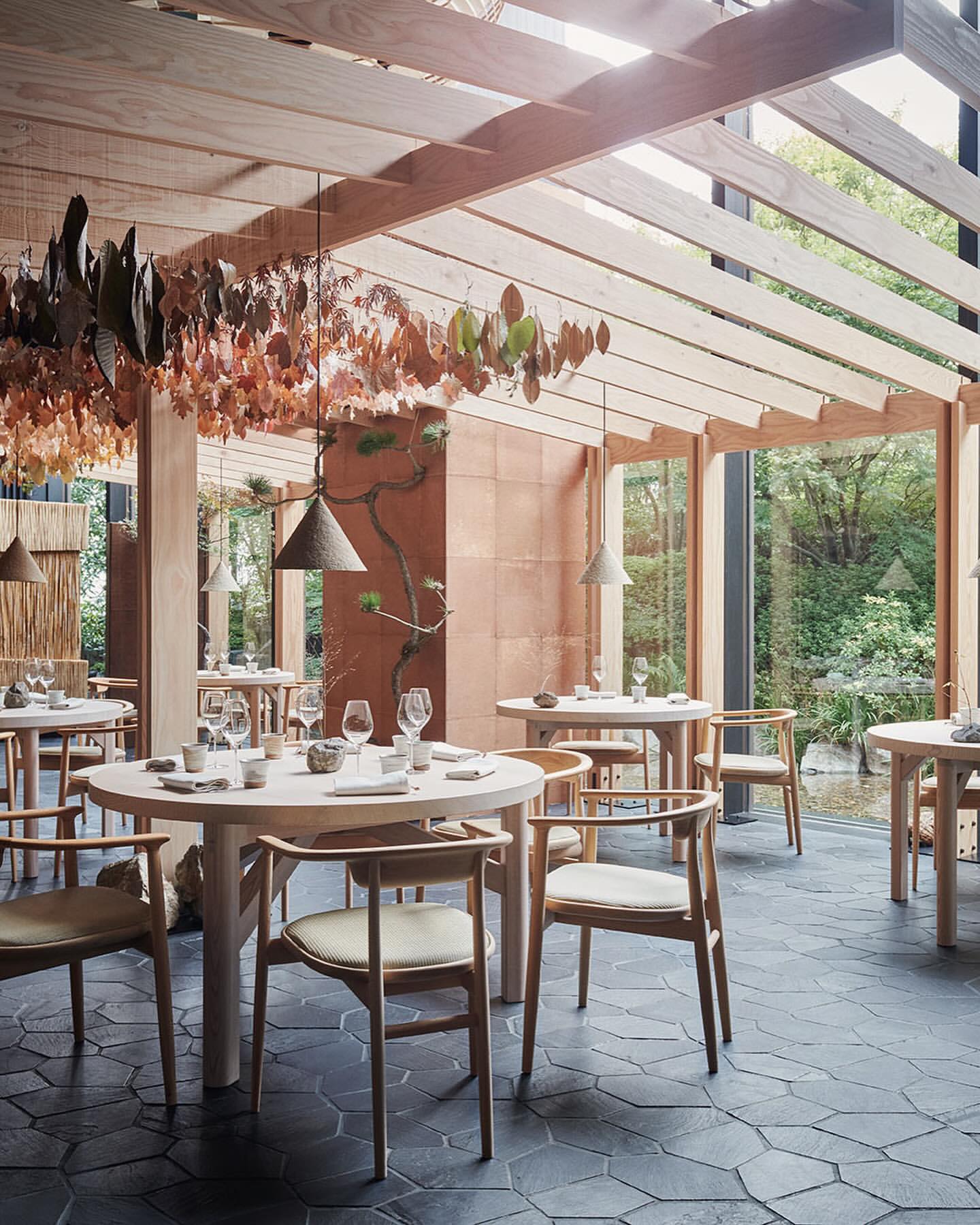 Restaurant interior featuring tables set for dining with modern wooden chairs, pendant lights, and a view of outdoor greenery.