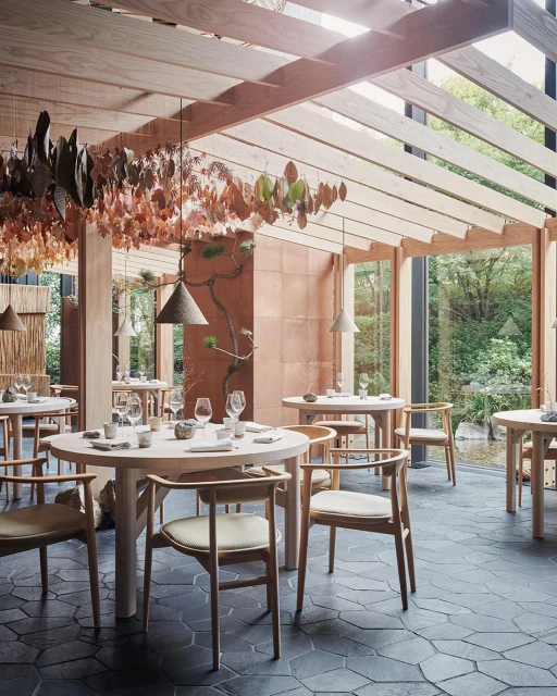 Restaurant interior featuring tables set for dining with modern wooden chairs, pendant lights, and a view of outdoor greenery.