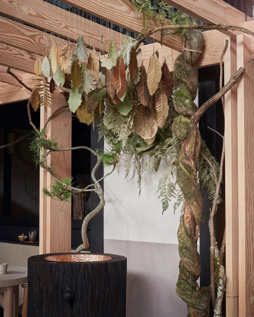 Interior shot of a modern sculptural wooden arbor featuring decorative metal leaves, a tree trunk water feature, and ambient lighting.