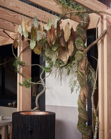 Interior shot of a modern sculptural wooden arbor featuring decorative metal leaves, a tree trunk water feature, and ambient lighting.