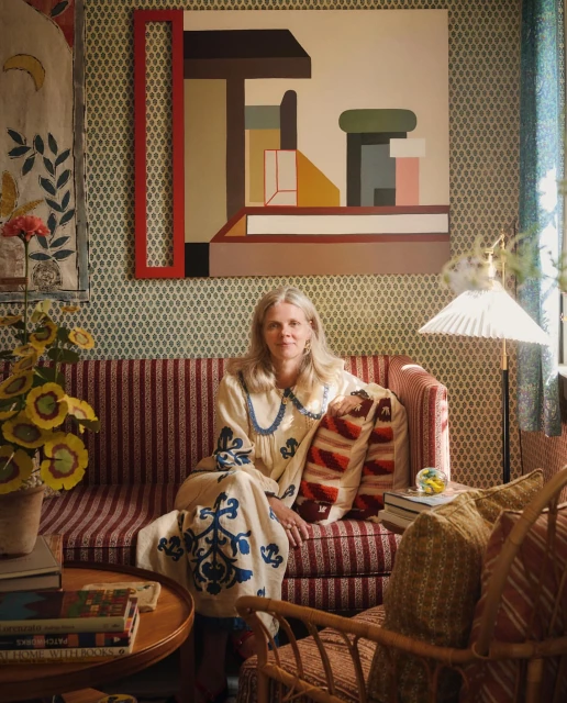 Blonde woman sits on a striped sofa with throw pillows in a room with floral wallpaper, geometric art, and a round table with books.