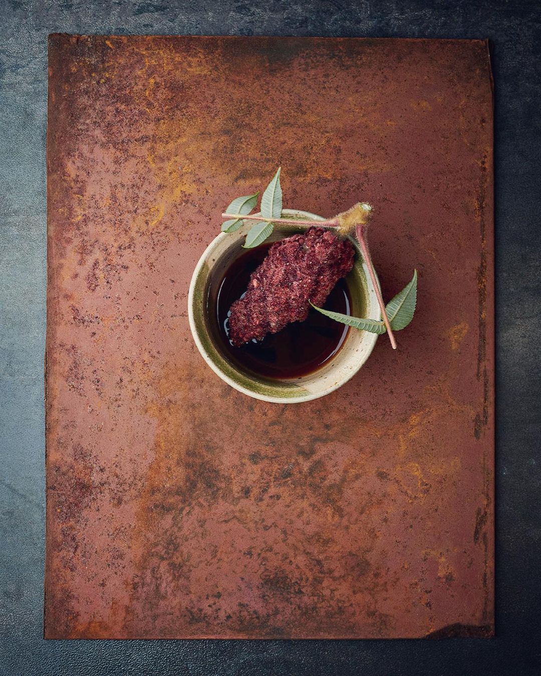 Overhead shot of a rustic wooden board with a bowl of dark liquid, garnished with a dark red food item and green leaves. 