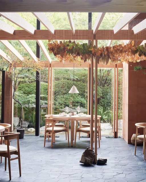 Interior of an elegant restaurant dining room with a light wood table and chairs, set against a backdrop of a lush outdoor garden.