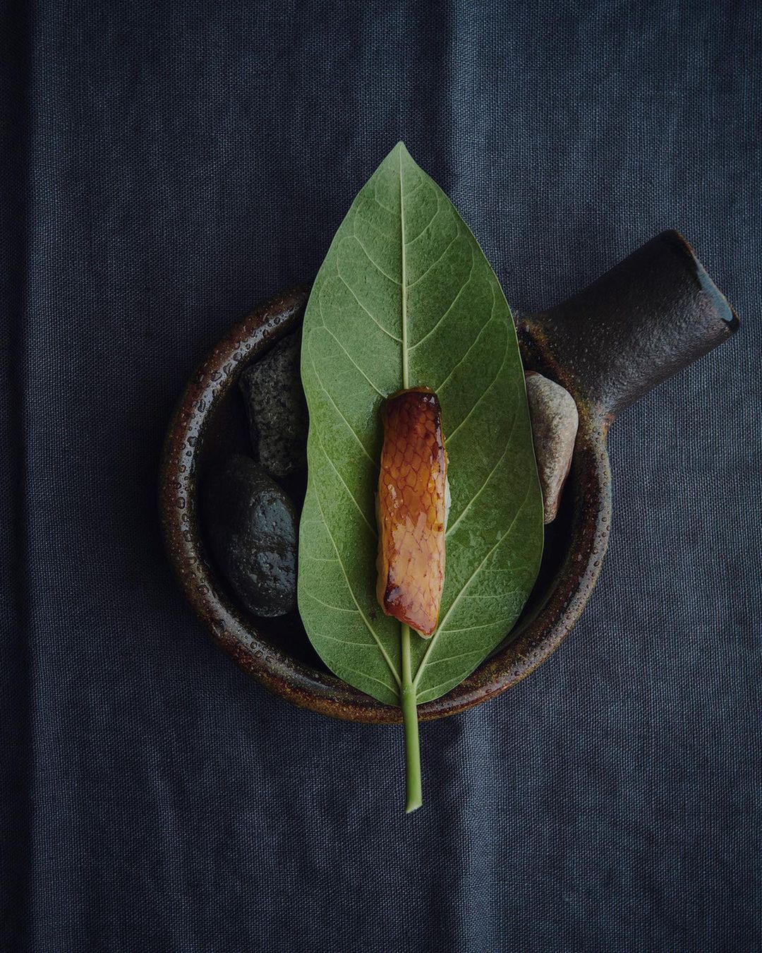 Overhead shot of glazed protein on a green leaf in a clay dish with pebbles on a gray textured surface. Food presentation.