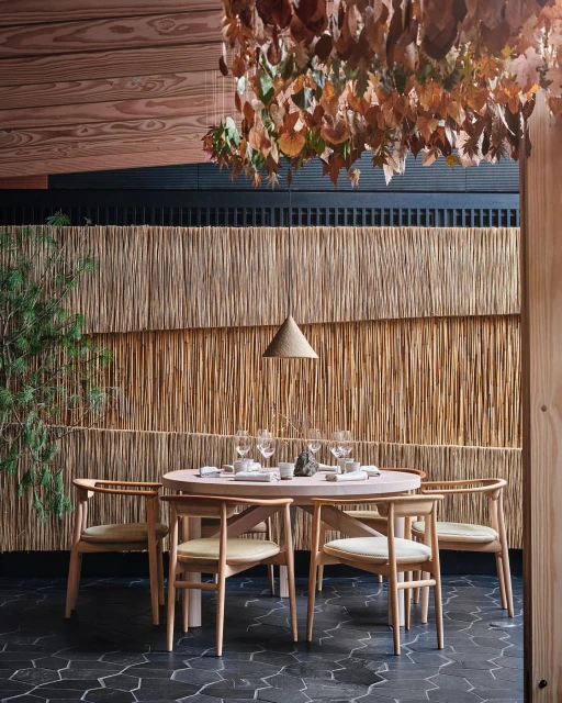 Restaurant dining area with round table set for a meal, surrounded by wooden chairs, with decorative lighting and a unique woven wall.
