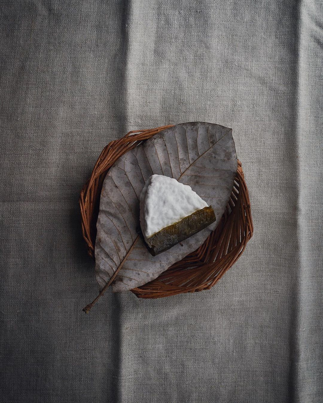 Overhead shot of a slice of cheese with a white rind on a brown leaf in a brown woven basket on a grey textured fabric.