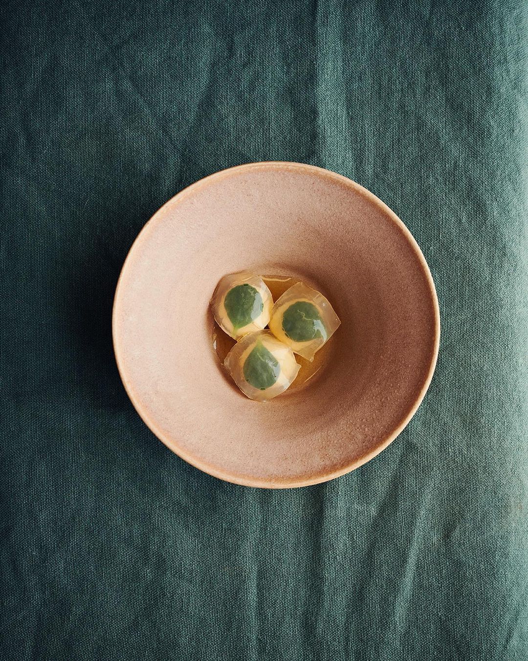 Overhead shot of a peach-colored bowl containing three translucent dumplings with green centers, resting on a dark green fabric surface.