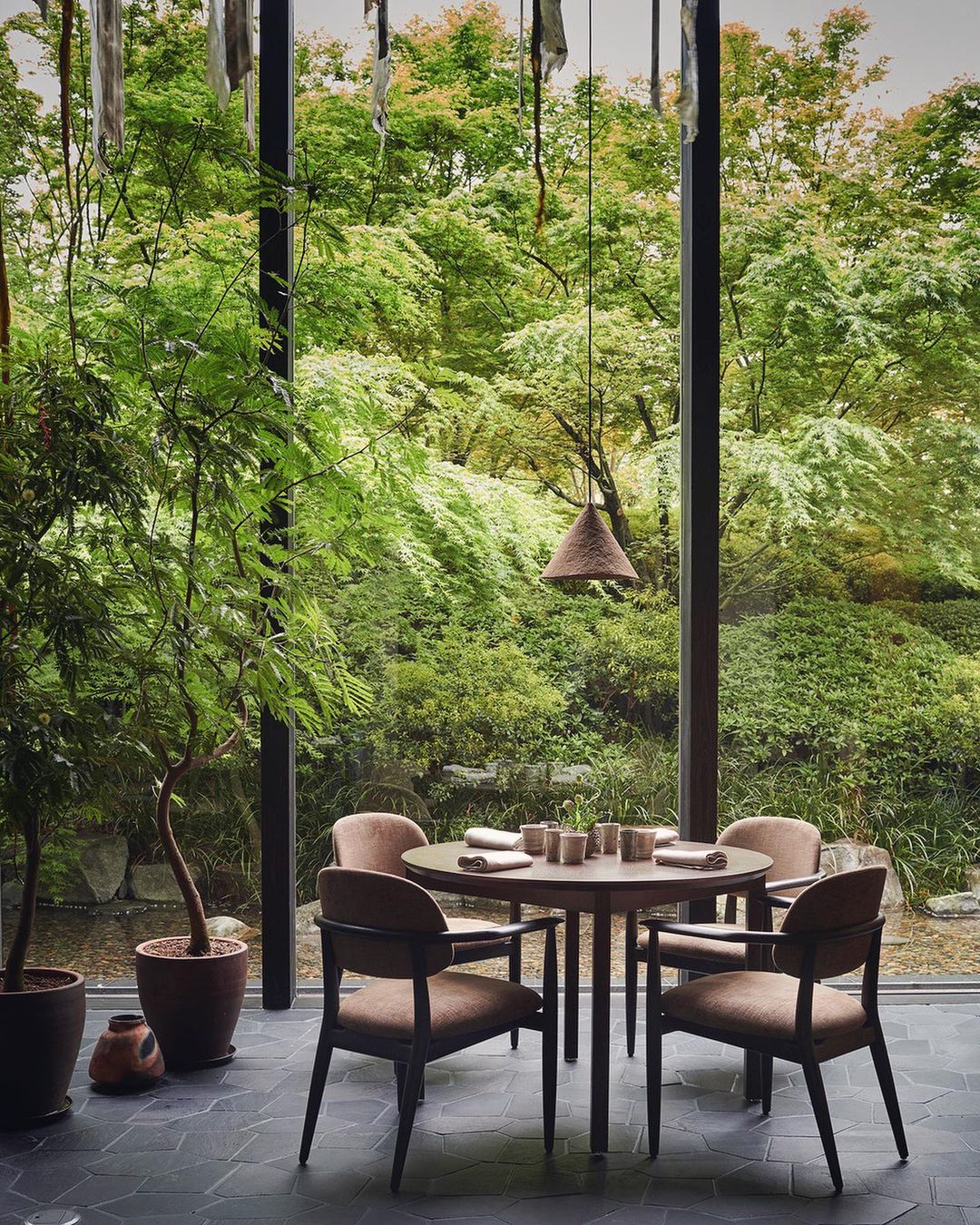 Restaurant interior features a round dining table with four chairs, set against a backdrop of lush green foliage visible through a window.