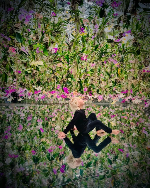 Young child in black velvet loungewear reclines barefoot on a reflective surface in a lush orchid garden. Indoor setting.