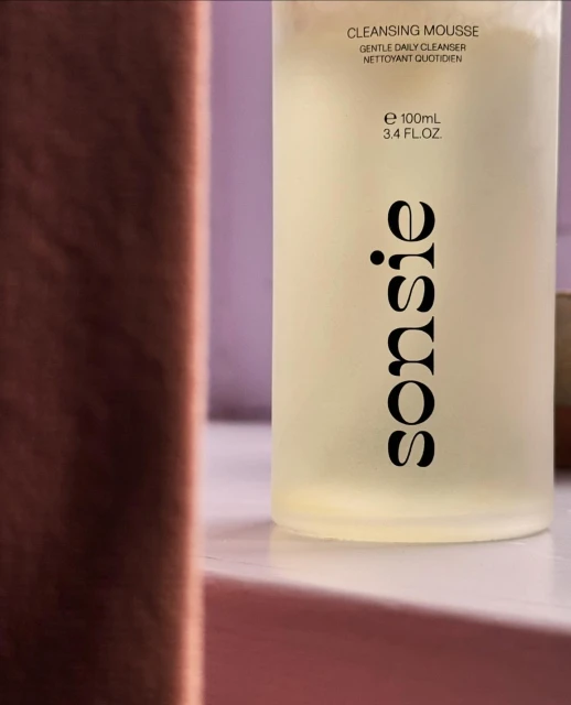A bottle of Sonsie cleansing mousse, featuring minimal design and product information, set against a soft pink and brown backdrop.