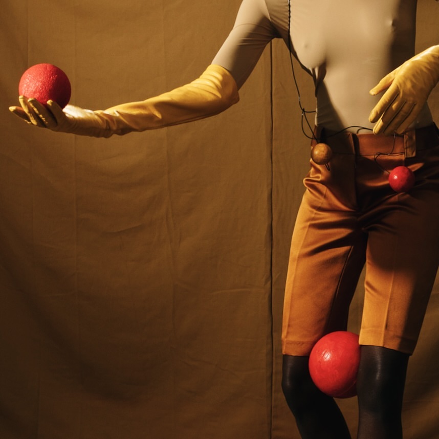 Person in a beige bodysuit and ochre shorts with red balls, against an ochre background. Creative fashion studio shot.