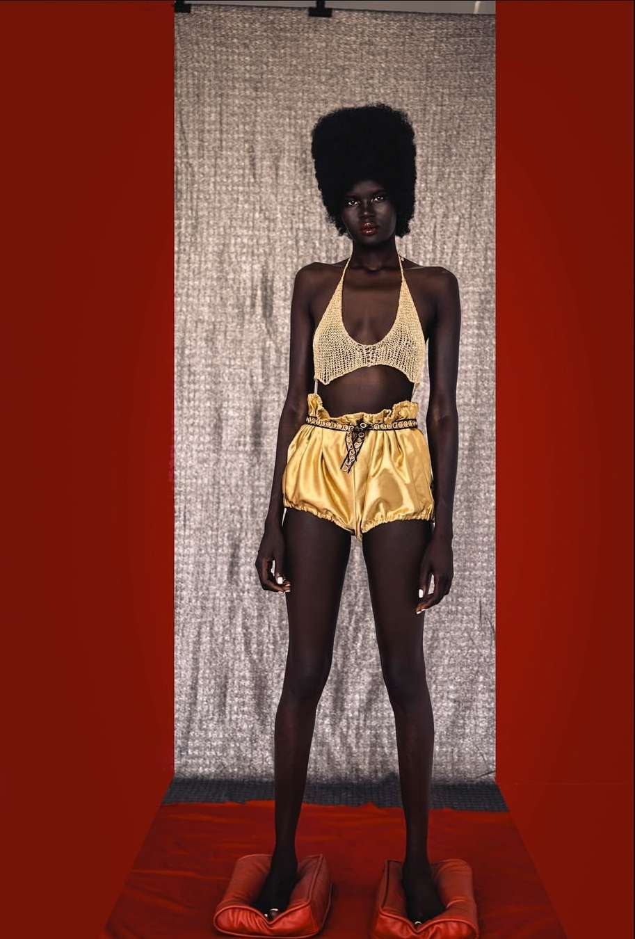 Dark-skinned model poses in a knit top and gold shorts against a textured backdrop, standing on red pillow-like platforms.