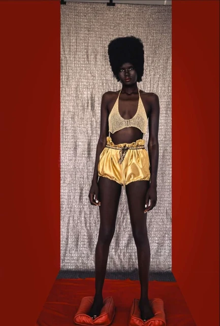 Dark-skinned model poses in a knit top and gold shorts against a textured backdrop, standing on red pillow-like platforms.