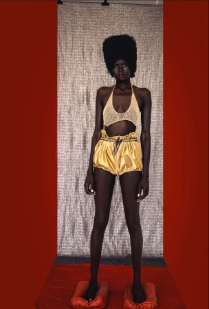 Dark-skinned model poses in a knit top and gold shorts against a textured backdrop, standing on red pillow-like platforms.