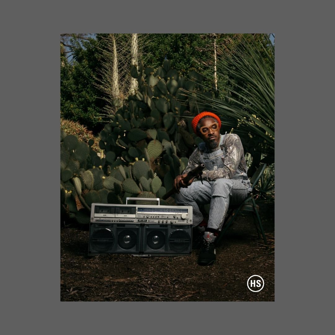 Adult man wearing a hat sitting outdoors with a boombox in front of a cactus plant, looking at camera with serious expression.