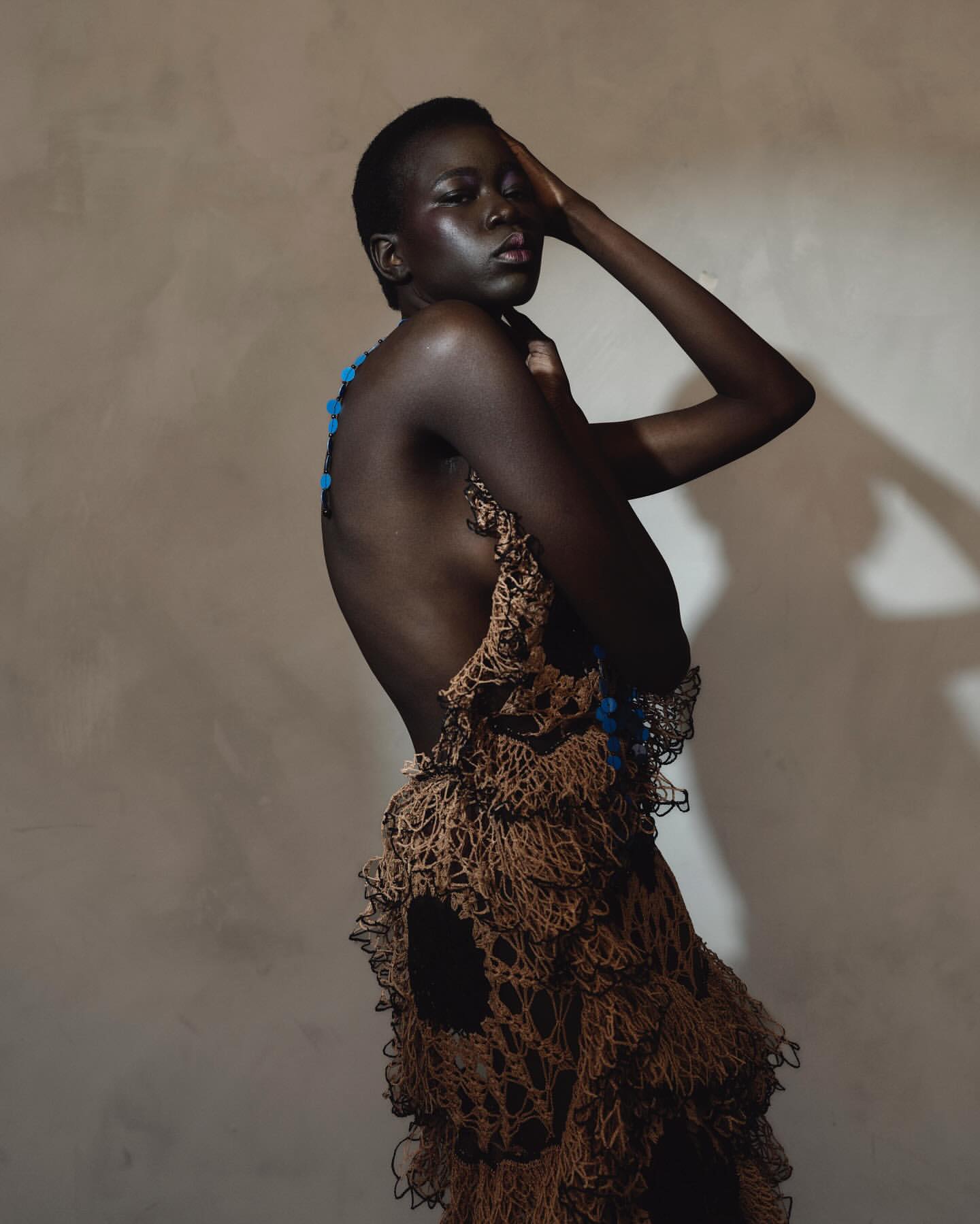 A young Black female model poses in a brown, netted, layered garment in front of a warm-toned backdrop in a studio fashion shot.