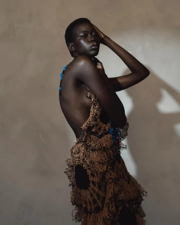 A young Black female model poses in a brown, netted, layered garment in front of a warm-toned backdrop in a studio fashion shot.