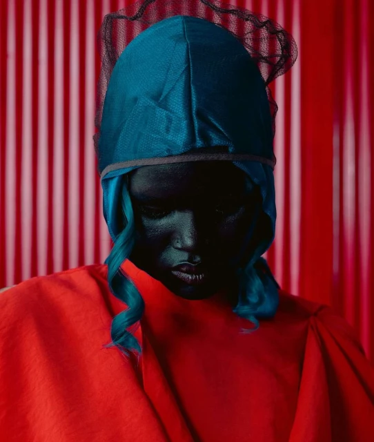 Close-up portrait of a woman with dark skin wearing a turquoise head covering and a red robe against a red and white striped backdrop.