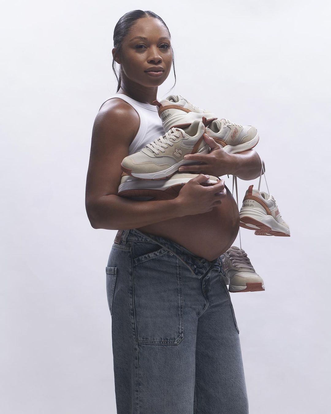 A pregnant woman holds a stack of sneakers in her arms, looking directly at the camera against a plain white background.