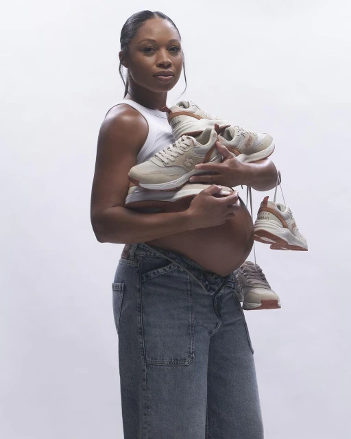 A pregnant woman holds a stack of sneakers in her arms, looking directly at the camera against a plain white background.