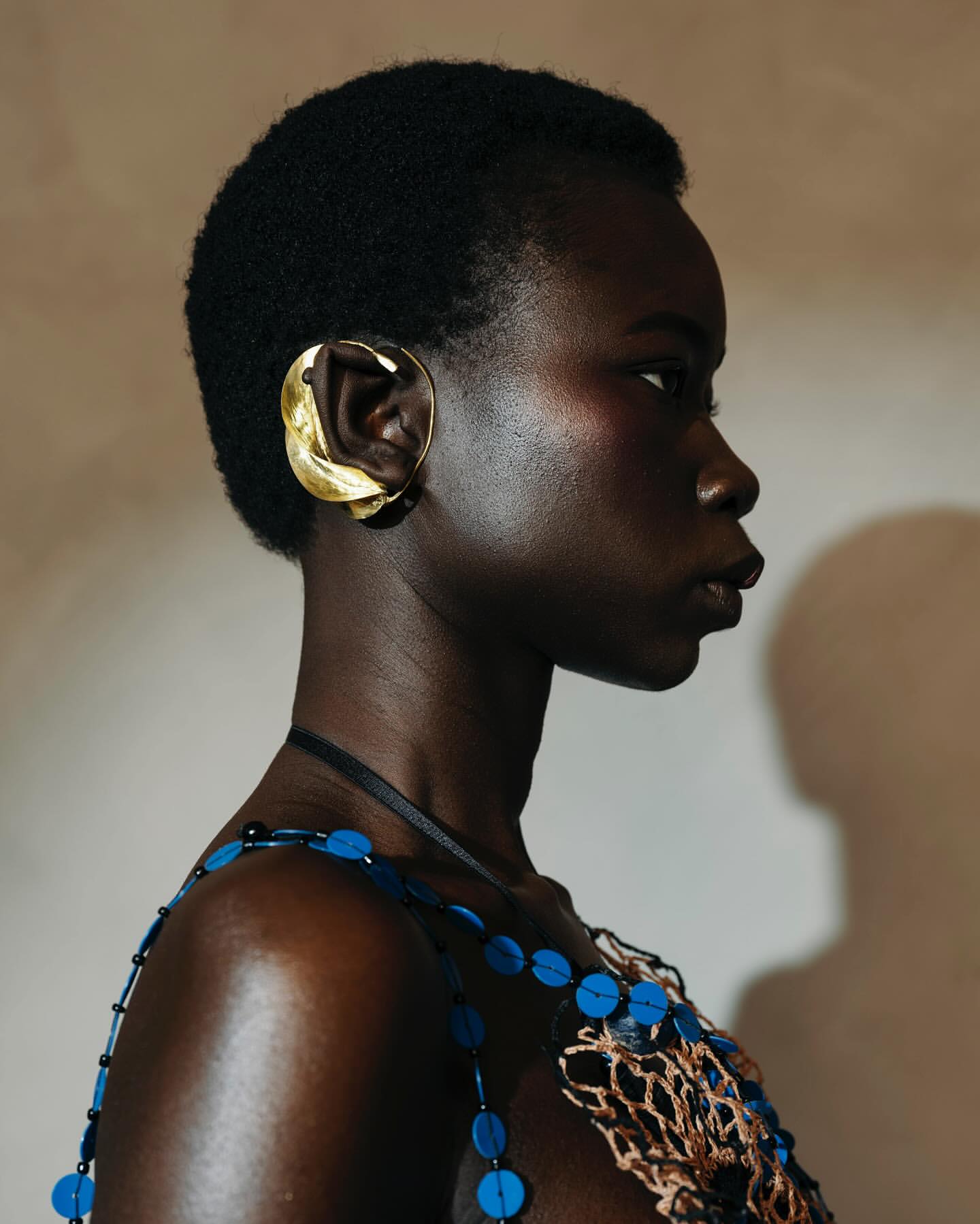 Close-up studio shot of a dark-skinned adult woman with short natural hair, adorned with a large gold sculptural ear cuff and beaded top.