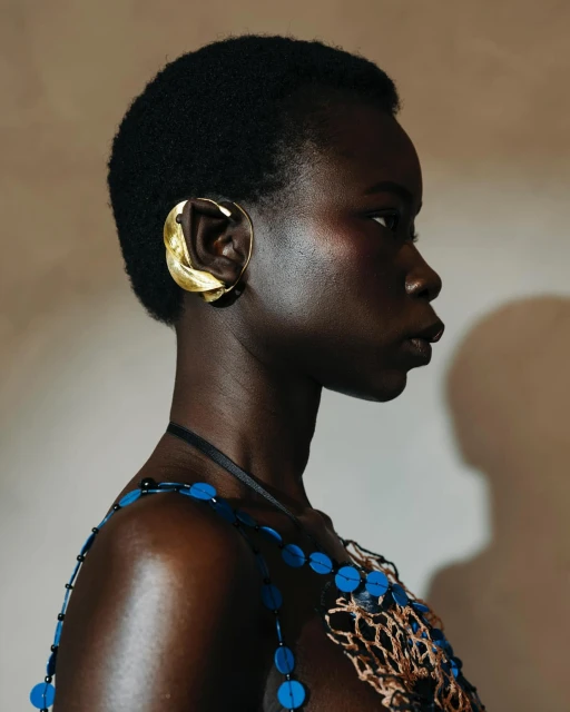 Close-up studio shot of a dark-skinned adult woman with short natural hair, adorned with a large gold sculptural ear cuff and beaded top.