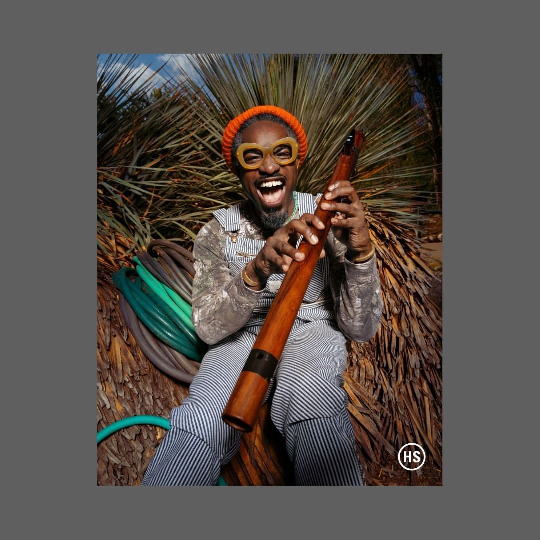 Enthusiastic Black man with an orange knit cap plays a didgeridoo outdoors, wearing overalls and round glasses, smiling with a desert backdrop.