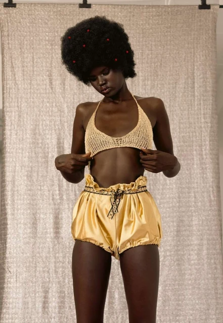 Young woman with dark skin and an afro hairstyle wearing a beige halter top and gold satin shorts poses against a neutral backdrop.