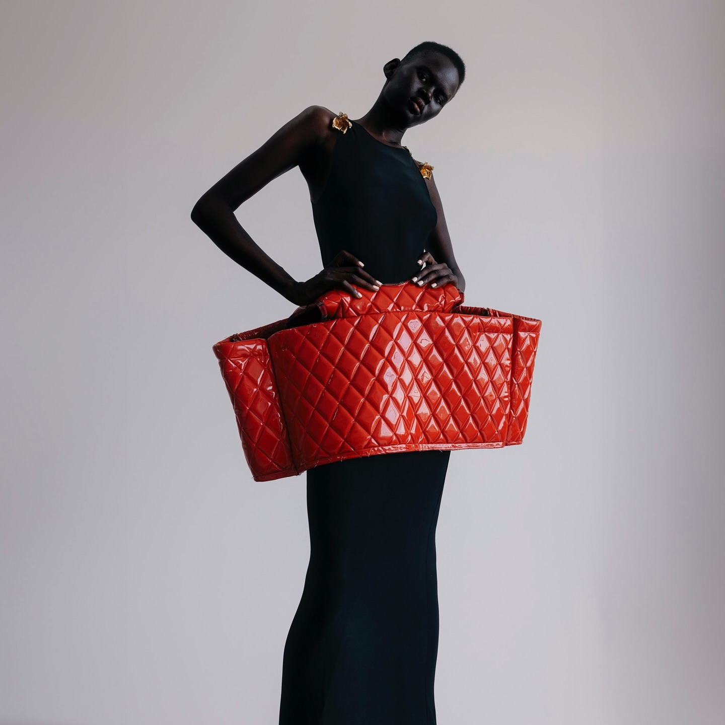 Fashionable woman in a black dress poses with a large, red, quilted bag-like structure around her waist in a studio setting.