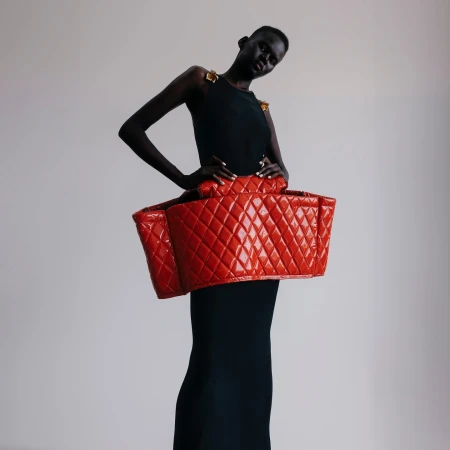 Fashionable woman in a black dress poses with a large, red, quilted bag-like structure around her waist in a studio setting.