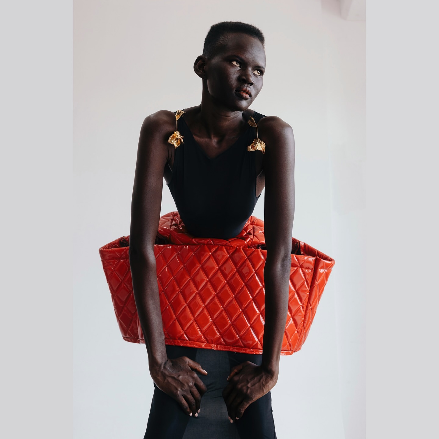 A statuesque Black woman in a black bodysuit poses against a bright background, carrying a large red quilted leather bag.