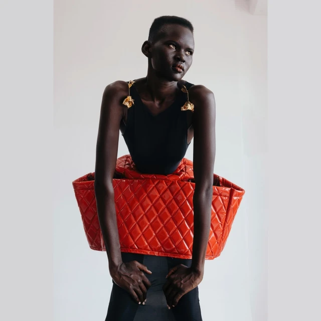 A statuesque Black woman in a black bodysuit poses against a bright background, carrying a large red quilted leather bag.