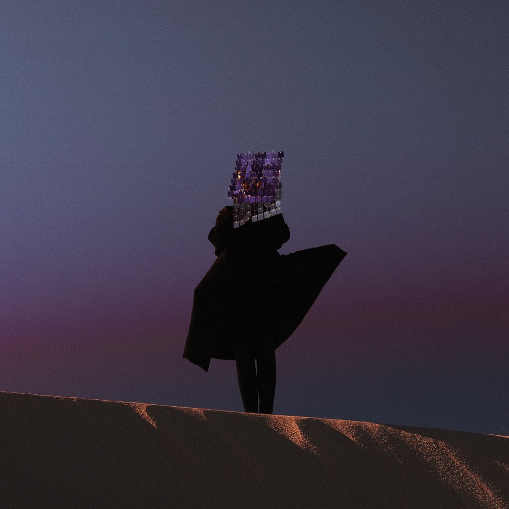 Person in a black coat stands on a sand dune with a cubical, reflective head covering at dusk with pink and purple sky.
