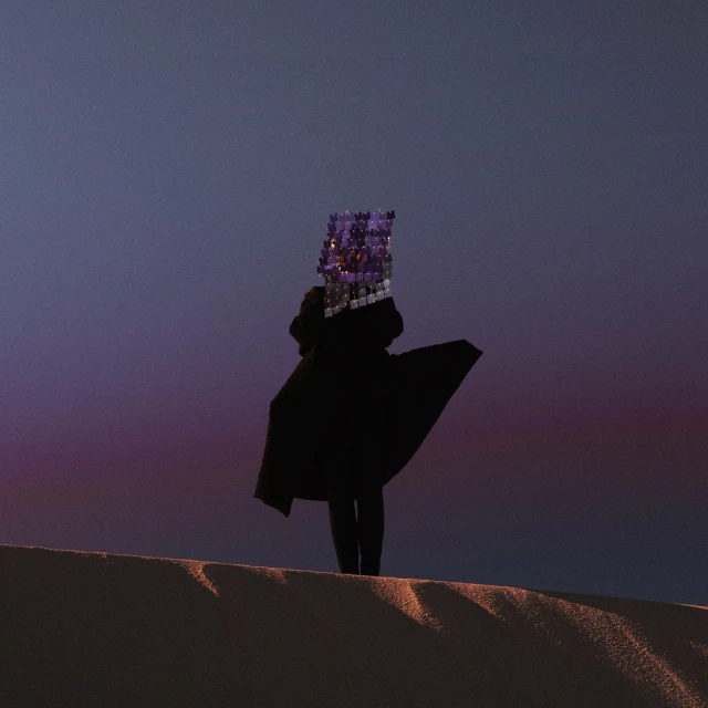Person in a black coat stands on a sand dune with a cubical, reflective head covering at dusk with pink and purple sky.