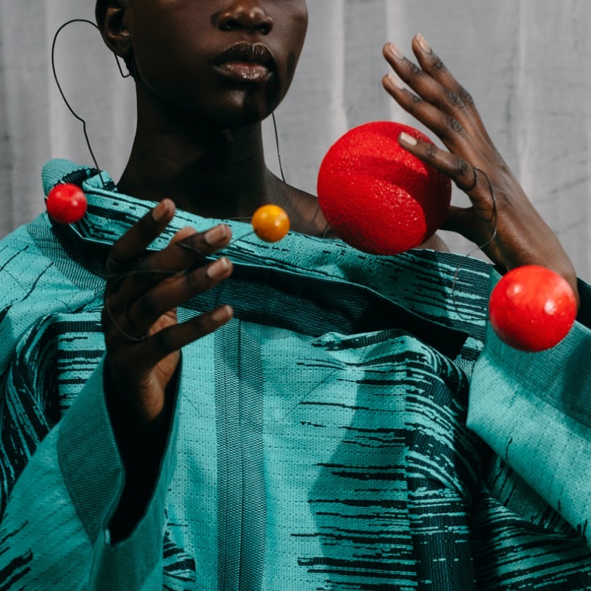 Striking studio shot of an African woman draped in a patterned teal textile holding suspended red and yellow spheres; stylish and artistic.