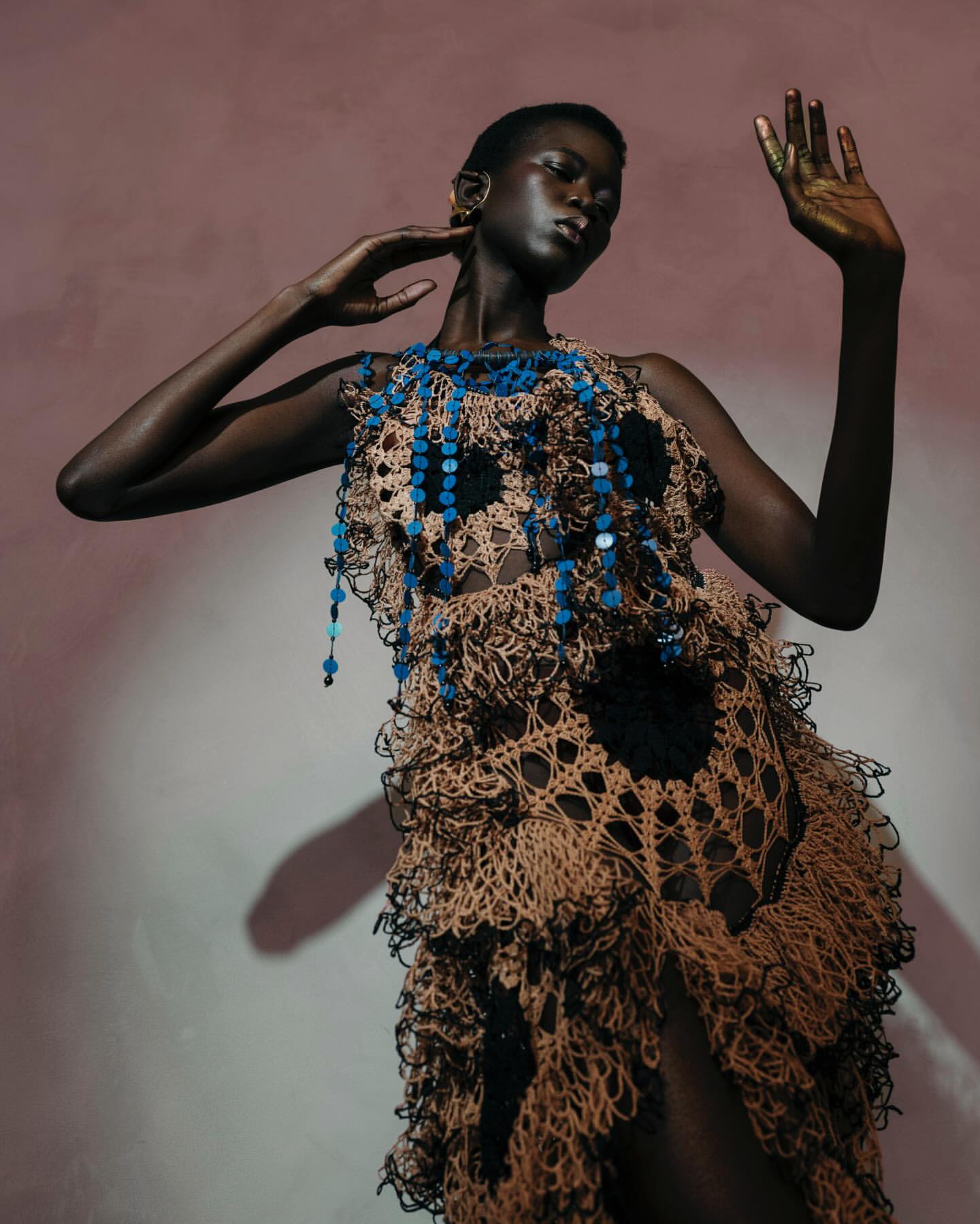 Fashion shot of a black woman wearing a crochet dress with blue beaded details, hands raised against a peach and grey background.