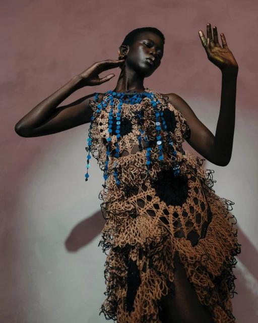 Fashion shot of a black woman wearing a crochet dress with blue beaded details, hands raised against a peach and grey background.