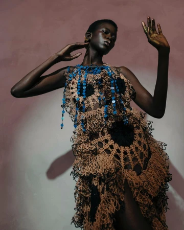 Fashion shot of a black woman wearing a crochet dress with blue beaded details, hands raised against a peach and grey background.