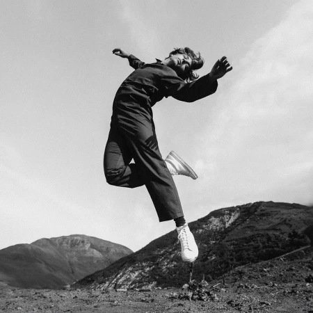 Black and white portrait of a person mid-air leaping against an outdoor landscape of rolling hills and sky.