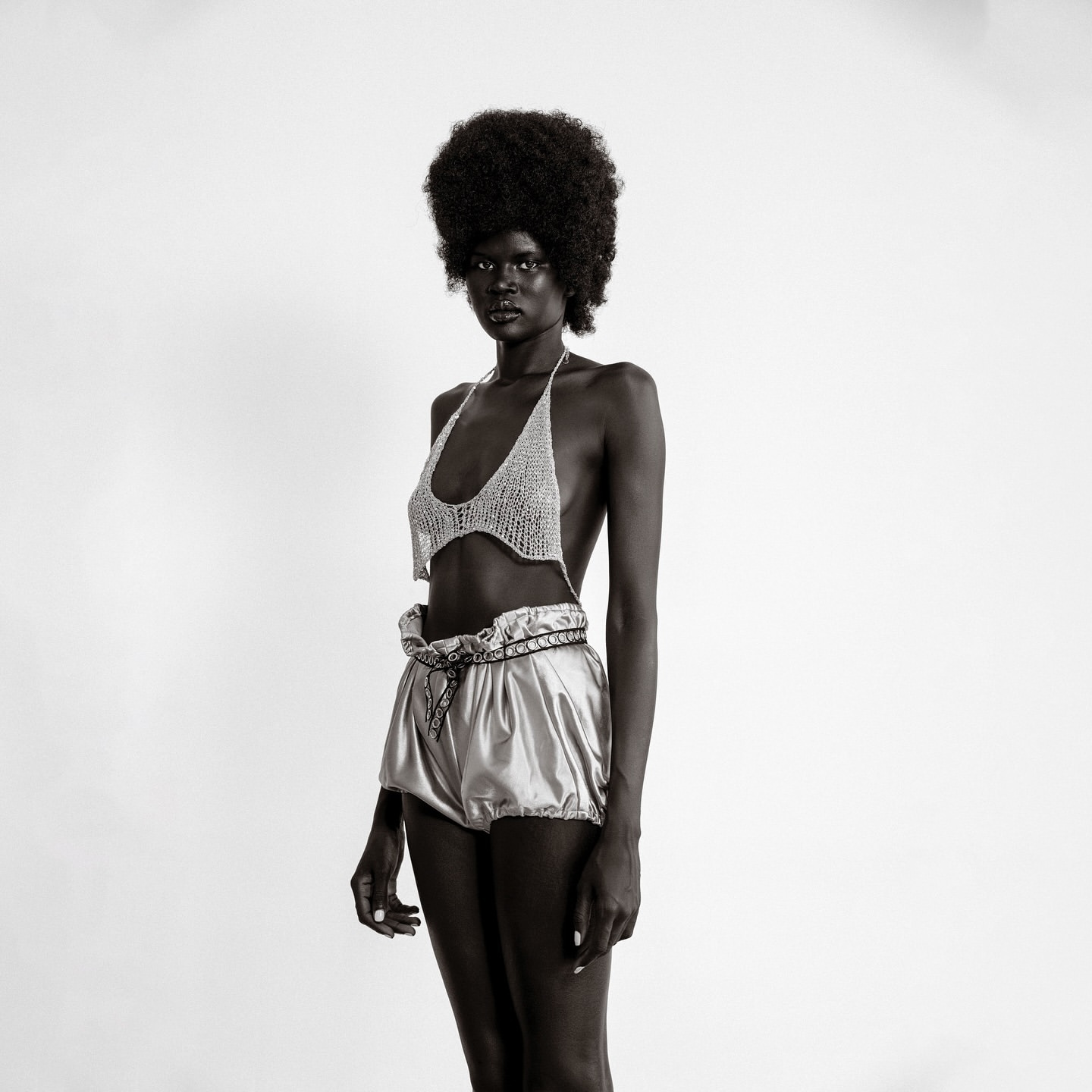 Black and white studio shot of a slender Black woman in a crocheted top and shiny shorts, showcasing fashion and style in simple setting.