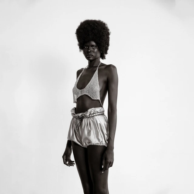Black and white studio shot of a slender Black woman in a crocheted top and shiny shorts, showcasing fashion and style in simple setting.