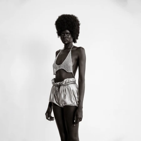 Black and white studio shot of a slender Black woman in a crocheted top and shiny shorts, showcasing fashion and style in simple setting.