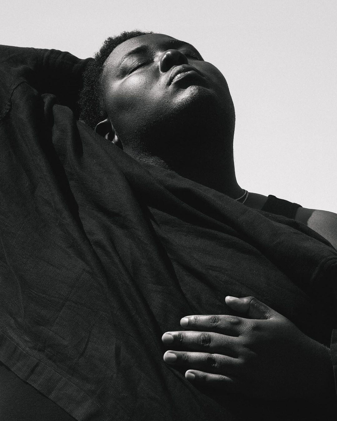Monochrome portrait of an adult, dark-skinned person with closed eyes, draped in dark fabric against a bright, neutral background.