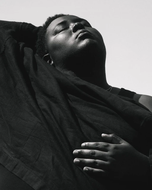 Monochrome portrait of an adult, dark-skinned person with closed eyes, draped in dark fabric against a bright, neutral background.