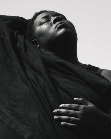Monochrome portrait of an adult, dark-skinned person with closed eyes, draped in dark fabric against a bright, neutral background.