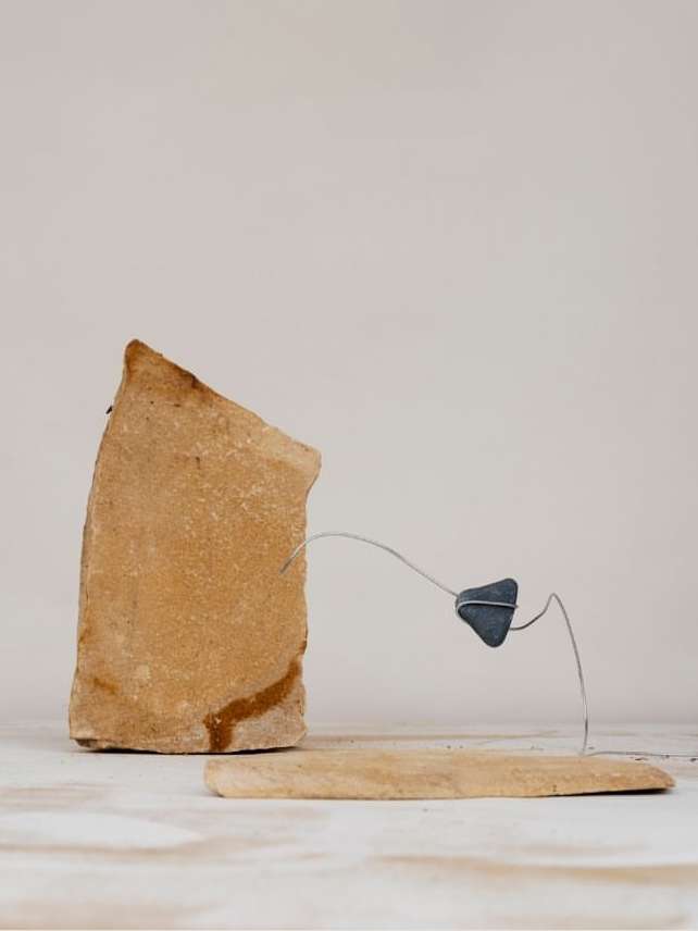 Still life featuring a tan rock formation connected by thin metal to a small dark rock piece, resting on a tan rock slab.