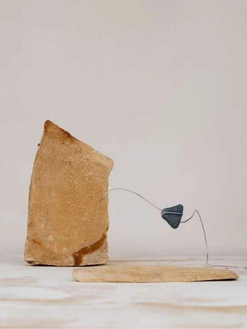 Still life featuring a tan rock formation connected by thin metal to a small dark rock piece, resting on a tan rock slab.